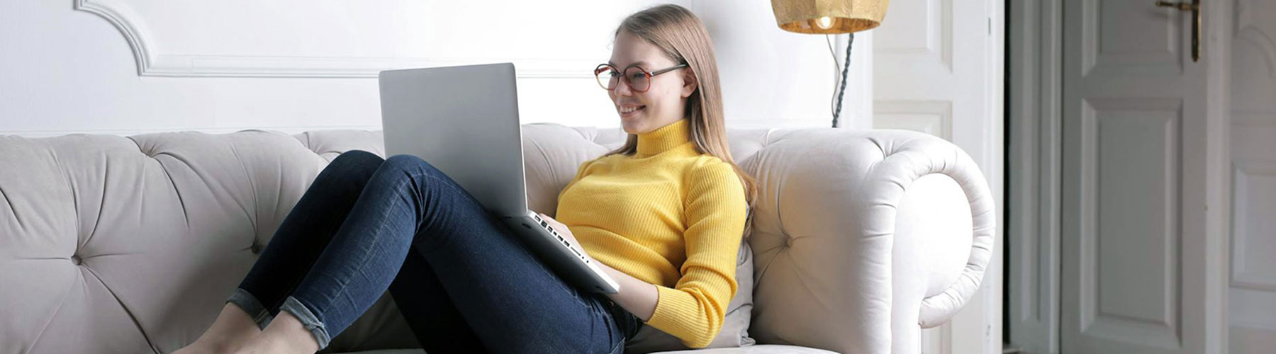 Smiling female in a yellow long sleeve shirt, jeans, and glasses happily working on a laptop while sitting on a couch in a living room