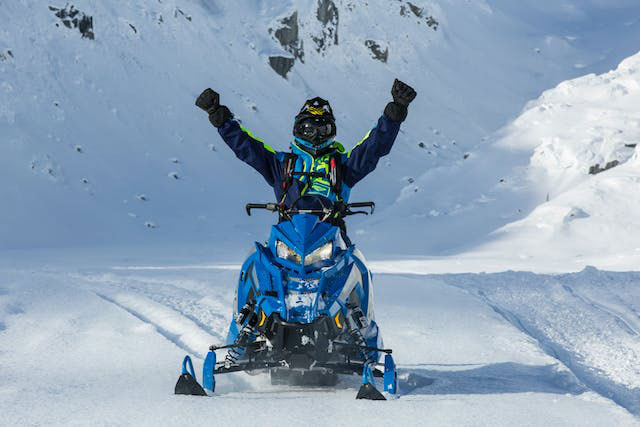 Guy sitting on a stationary snowmobile in the wilderness with his arms up cheering