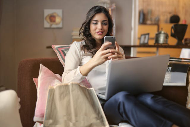 Smiling female sitting on a couch in a living room using a smartphone and a laptop