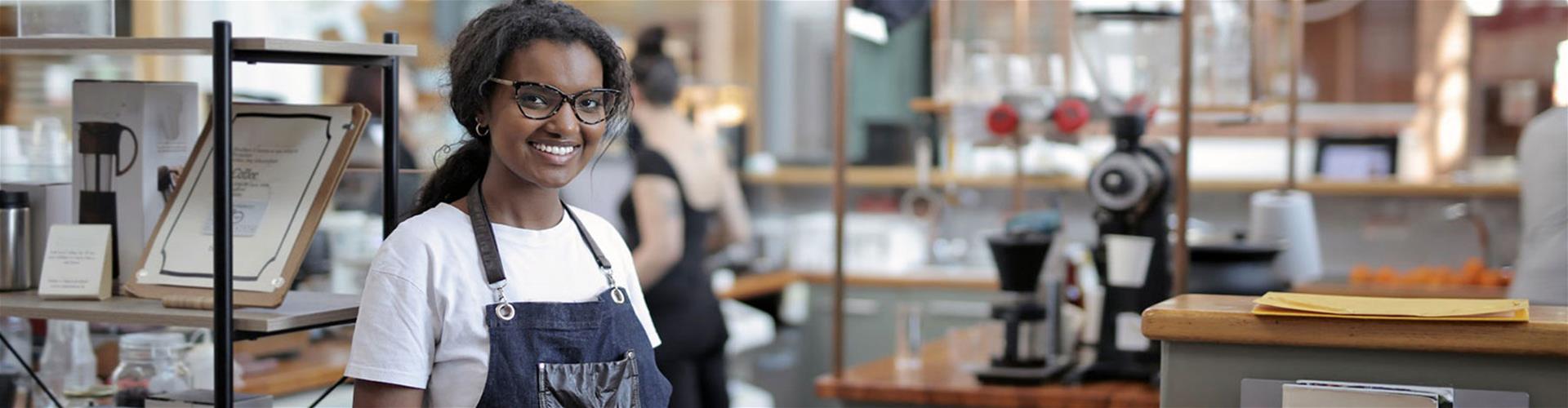 Happy woman wearing an apron standing by shelves in café Happy woman wearing an apron standing by shelves in café