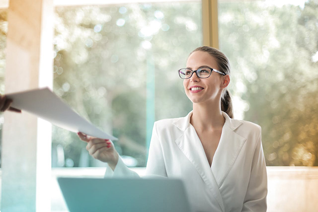 Smiling business woman at her desk in an office being handed a small stack of documents Smiling business woman at her desk in an office being handed a small stack of documents