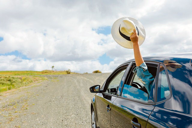 Navy blue sedan driving down a dirt road in the country on a partly cloudy day the windows are rolled down and a back passage is holding a hat and sticking their hand out of the window