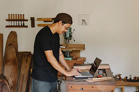 Concentrated ethnic male browsing laptop in woodworker workshop