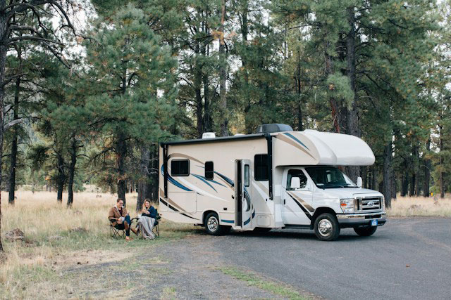 Couple sitting outside their RV in chairs at a campsite with conifer trees behind them