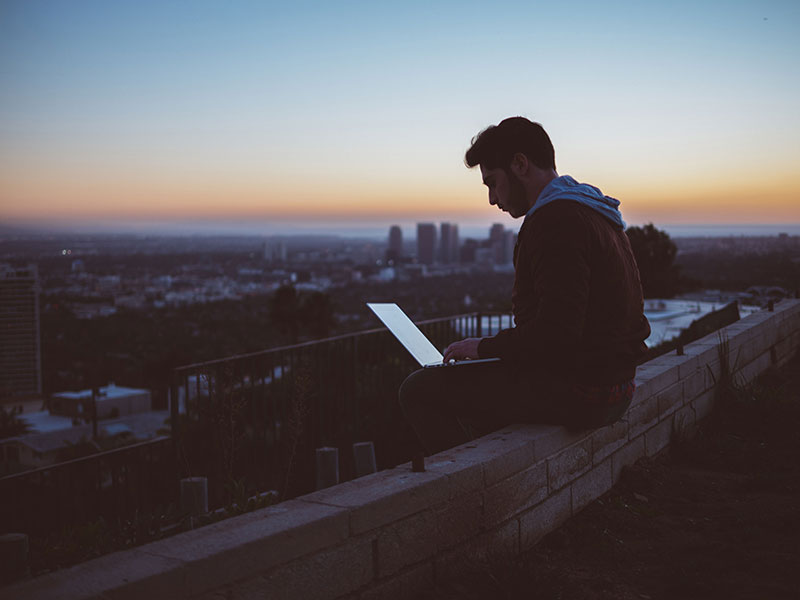 Man sitting on city roof during sunset on his computer
