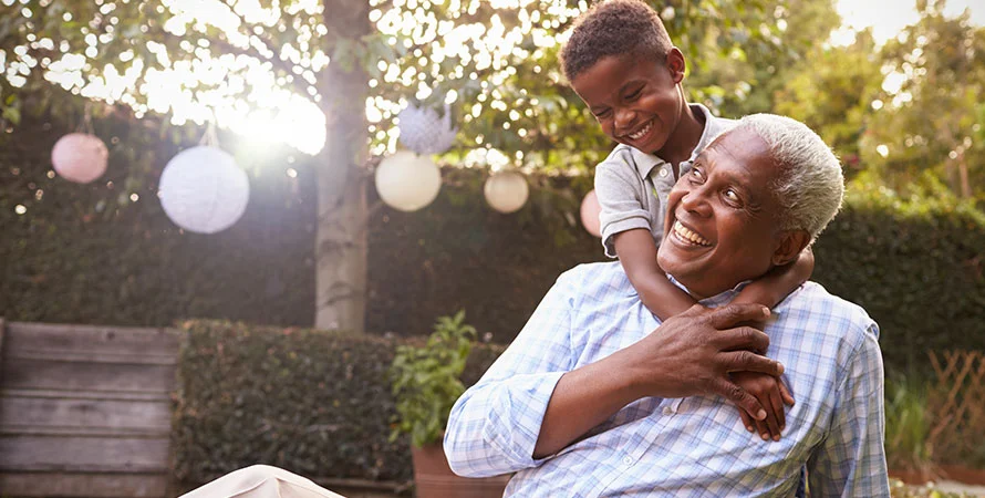 Smiling grandfather playing with grandson in a sunlit backyard with hanging lanterns.