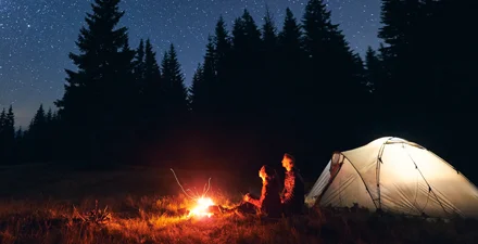 Two people at fire under the stars next to tent with light on.