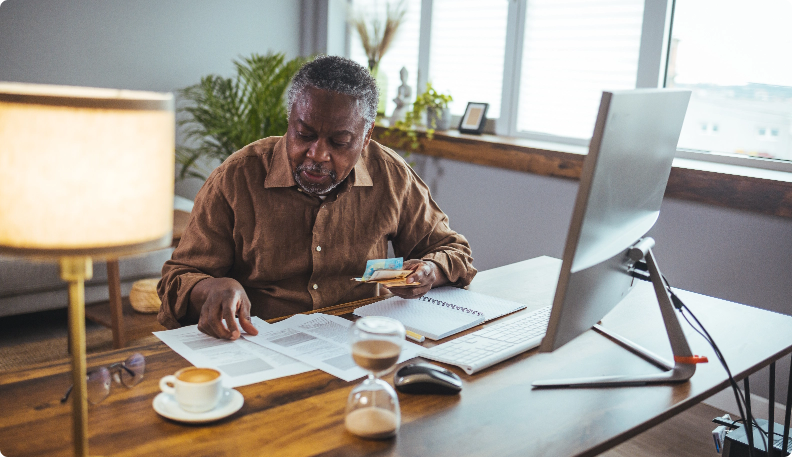 Elder man sitting at his wood computer table with coffee and paying bills.