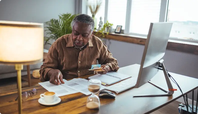 Elder man sitting at his wood computer table with coffee and paying bills.