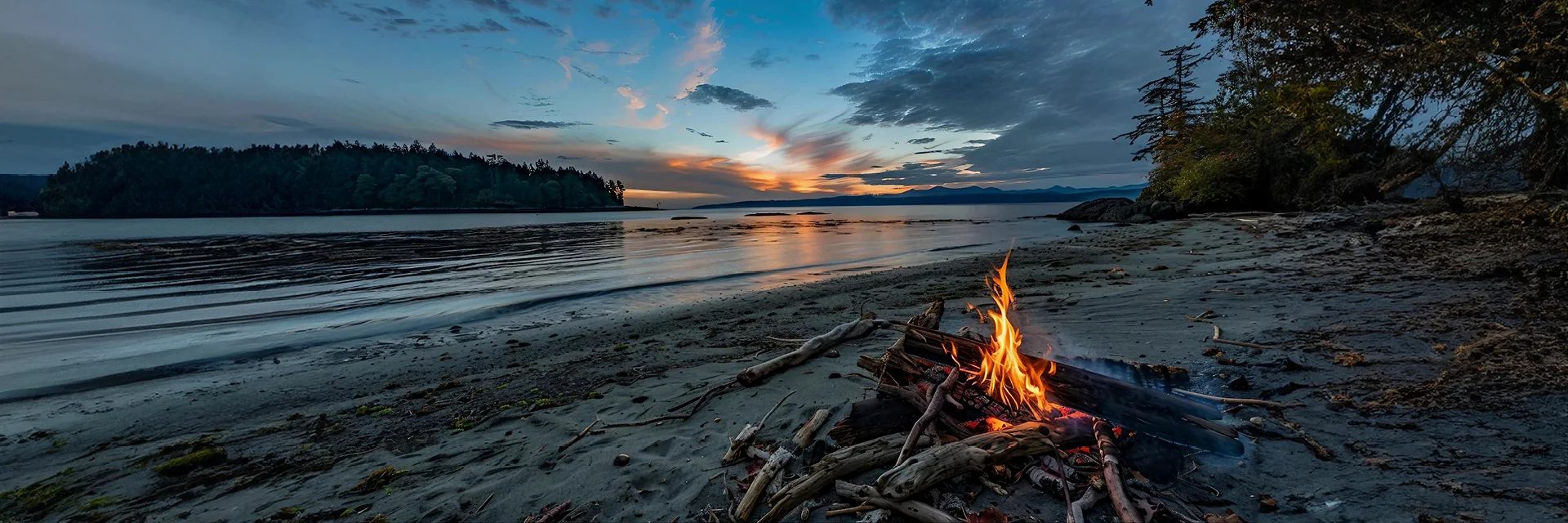 Campfire on a beach overlooking an island and lake at sunset before dark.