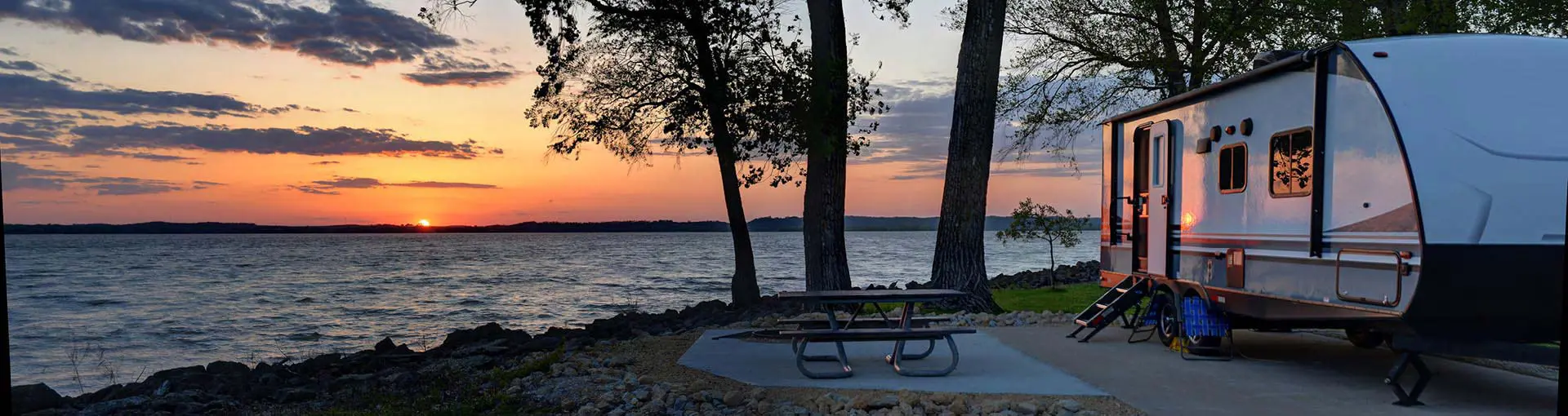 Travel trailer parked by a lake at sunset with trees and a picnic table nearby.