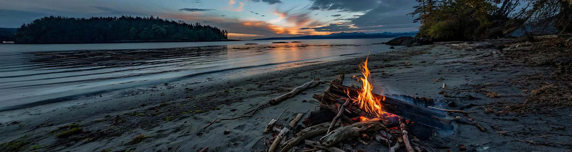 Campfire burning on a sandy beach at sunset with calm water and forested island in the distance.