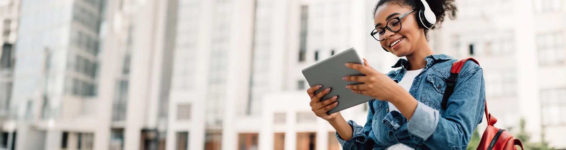 Smiling young woman with headphones using a tablet outdoors in front of tall city buildings.