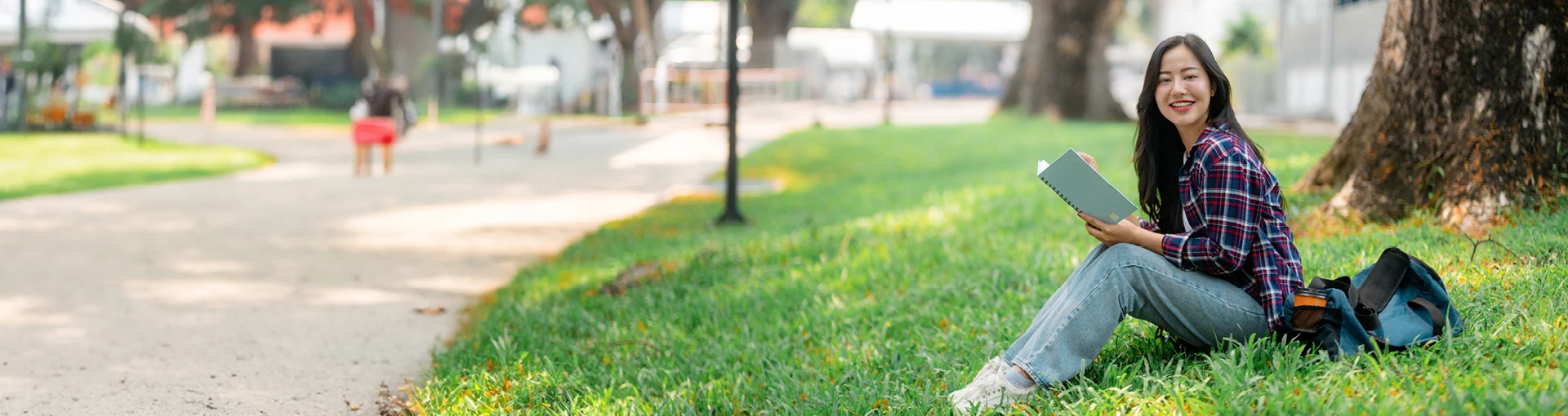 Female college student sitting on the grass in local park reading next to book bag.