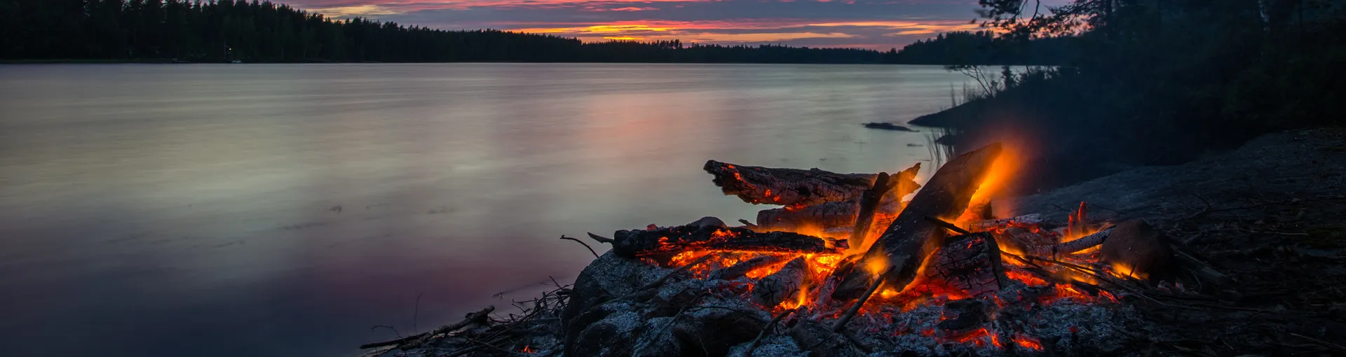 Fire smoldering next to a calm lake at sunset with hills in the distance.