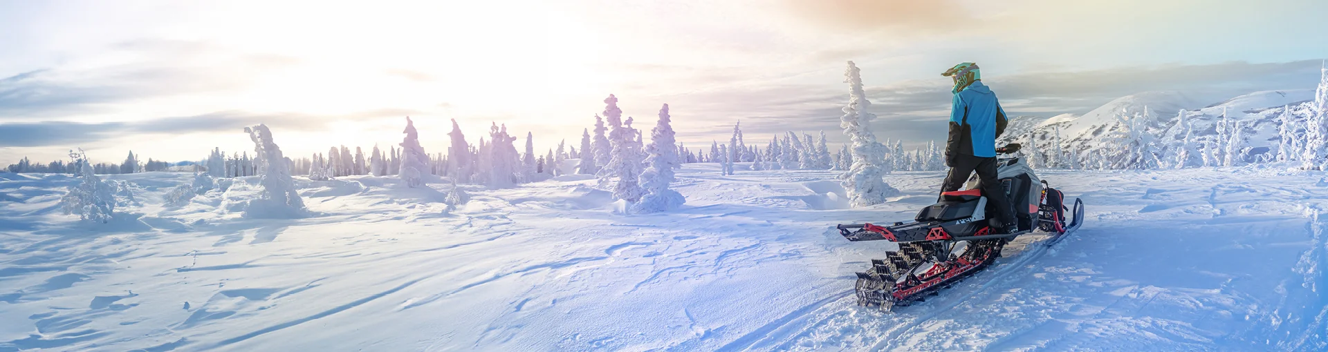 Man on snowmobile at sunrise in the winter on top of a ridge with trees and hills around.