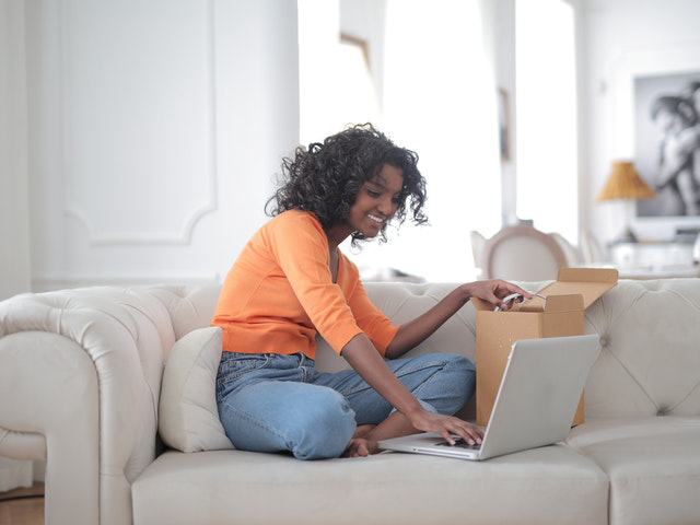 Cheerful woman sitting on couch opening box and using laptop