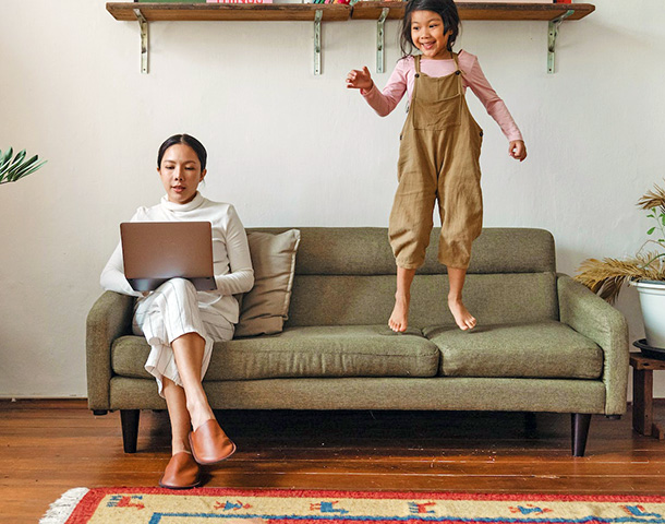 focused mother working on laptop near daughter jumping on couch