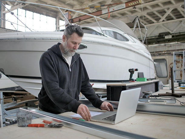 worker using laptop while working with metal parts near boat in workshop