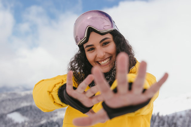 Woman in yellow jacket and ski goggles outside in the snow waving at camera