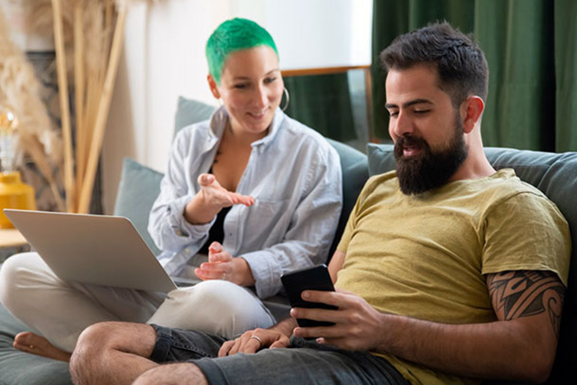 man using a phone sitting beside woman using a laptop on a couch at home man using a phone sitting beside woman using a laptop on a couch at home