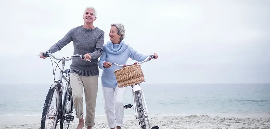An older couple smiling and walking bikes on beachfront.