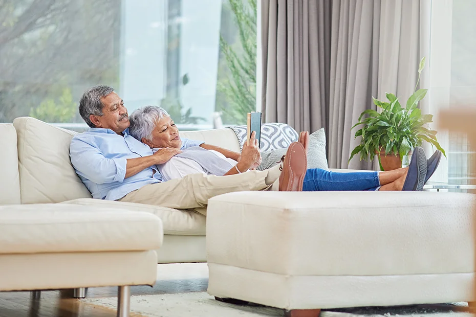 Older couple sitting together on a light couch in well lit living room reading.